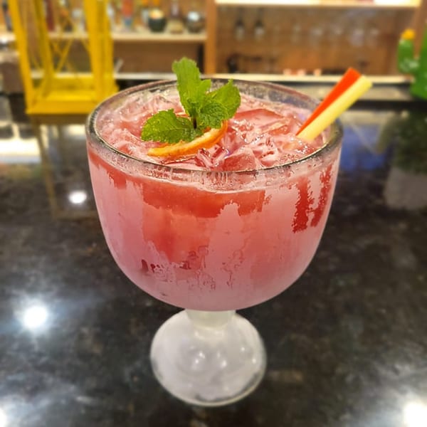 A large, frosty red margarita with ice, mint, and an orange slice, on a dark bar.